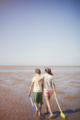 Bruder und Schwester mit Schaufeln, die sich umarmen und im nassen Sand am sonnigen Sommerstrand unter blauem Himmel laufen - HOXF02194