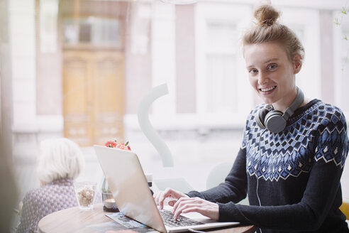 Porträt einer lächelnden jungen Frau, die in einem Caféfenster einen Laptop benutzt - HOXF02200
