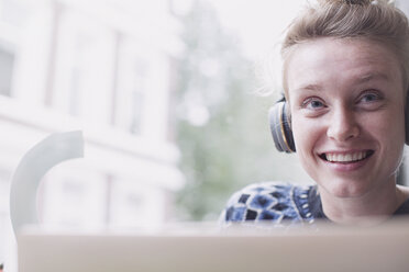 Portrait smiling young woman wearing headphones at laptop in cafe window - HOXF02205