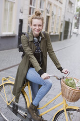 Porträt einer lächelnden jungen Frau mit Kopfhörern, die mit einem Korb voller Produkte Fahrrad fährt - HOXF02206