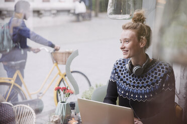 Smiling young woman with headphones using laptop in urban cafe window - HOXF02219