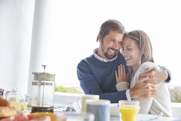 Lächelndes, zärtliches Paar, das sich am Frühstückstisch auf der Terrasse umarmt - HOXF02274