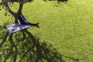 Gelassener Geschäftsmann entspannt sich auf einer Decke unter einem Baum in einem sonnigen Park - HOXF02294