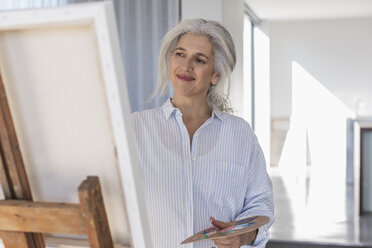 Smiling mature woman with palette painting at canvas on easel - HOXF02300