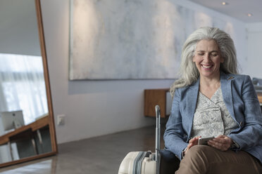 Smiling mature woman with suitcase texting with cell phone - HOXF02332