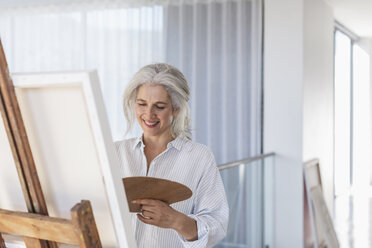 Smiling mature woman with palette painting at canvas on easel - HOXF02370