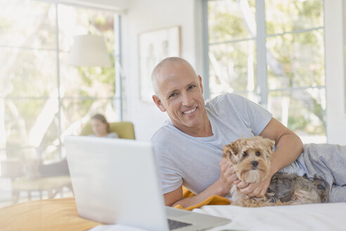 Porträt lächelnder Mann, der einen Hund am Laptop auf dem Bett streichelt - HOXF02402