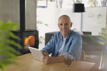 Portrait smiling man using digital tablet and drinking tea near wood stove fireplace - HOXF02421