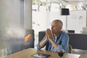 Pensive man with digital tablet drinking coffee and looking away near wood burning stove fireplace - HOXF02423