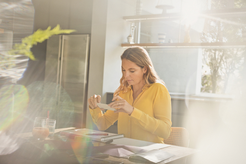 Frau mit Fotohandy fotografiert Kunst am sonnigen Küchentisch, lizenzfreies Stockfoto