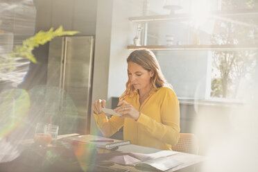 Woman with camera phone photographing art at sunny kitchen table - HOXF02433