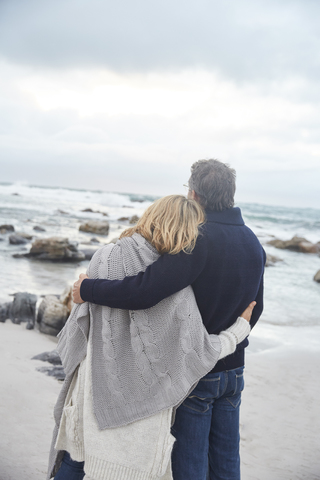 Gelassenes, zärtliches Paar, das sich am Winterstrand umarmt und auf das Meer schaut, lizenzfreies Stockfoto