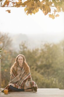 Serene woman wrapped in blanket meditating on autumn patio - HOXF02692