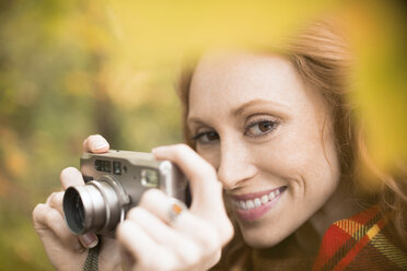 Nahaufnahme einer lächelnden Frau mit Digitalkamera inmitten von Herbstblättern - HOXF02710
