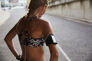 Fitte Läuferin im Sport-BH mit mp3-Player-Armband und Kopfhörern, die sich auf einer städtischen Straße ausruht - HOXF02720