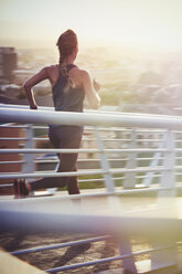Female runner running on sunny urban footbridge at sunrise - HOXF02722