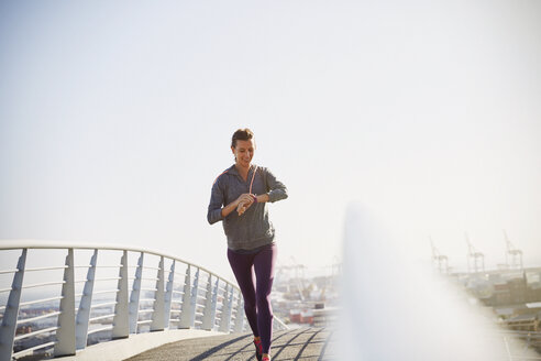 Lächelnde Läuferin, die auf einer sonnigen Fußgängerbrücke ihren Smartwatch-Fitness-Tracker überprüft - HOXF02750