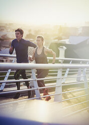 Runner couple running on sunny urban footbridge at sunrise - HOXF02779