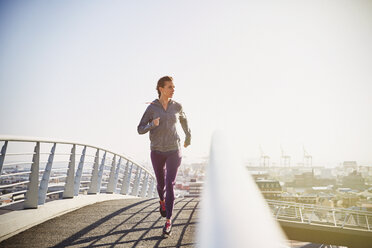 Female runner running on sunny urban footbridge at sunrise - HOXF02796