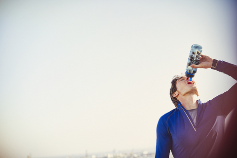 Durstiger männlicher Läufer trinkt Wasser aus einer Wasserflasche unter blauem Himmel, lizenzfreies Stockfoto