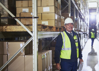 Portrait smiling manager with clipboard next to shelf in distribution warehouse - HOXF02893