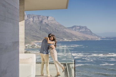 Paar, das sich auf dem sonnigen Luxusbalkon mit Blick auf das Meer und die Berge umarmt - HOXF02923