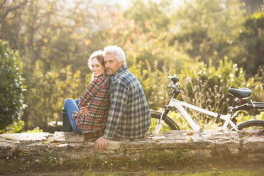 Portrait affectionate couple with bicycle resting on stone wall in autumn park - HOXF03102