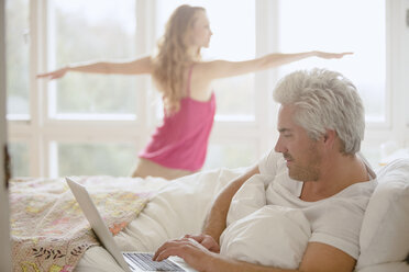 Frau übt Yoga-Krieger 2 Pose im Schlafzimmer mit Mann mit Laptop im Bett - HOXF03118