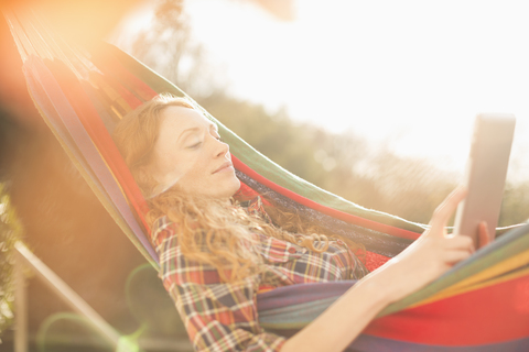 Frau entspannt sich in sonniger Hängematte mit digitalem Tablet, lizenzfreies Stockfoto