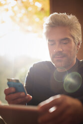 Mann mit digitalem Tablet und Handy im sonnigen Fenster - HOXF03123