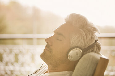 Gelassener Mann mit Kopfhörern beim Musikhören auf einer sonnigen Veranda - HOXF03130