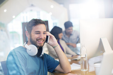Lächelnder kreativer Geschäftsmann mit Kopfhörern, der im Büro mit seinem Handy telefoniert - HOXF03155