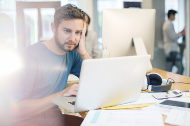 Seriöser kreativer Geschäftsmann mit Laptop und Kopf in den Händen im Büro - HOXF03162