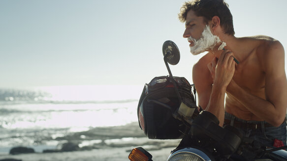 Junger Mann auf Motorrad rasiert Bart in der Nähe des Meeres - HOXF03317