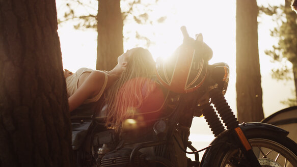 Junge Frau auf dem Motorrad liegend unter sonnigen Bäumen - HOXF03320