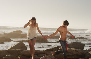 Young couple holding hands and walking on ocean rocks - HOXF03326