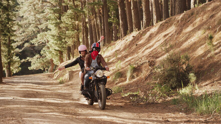 Exuberant young woman riding motorcycle on dirt road in woods - HOXF03334