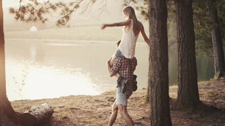 Young man carrying woman on shoulders at lakeside - HOXF03335