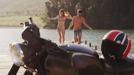 Junges Paar, das auf einem Steg am Seeufer in Richtung Motorrad läuft - HOXF03343