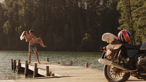 Young man carrying young woman on lakeside dock near motorcycle stock photo