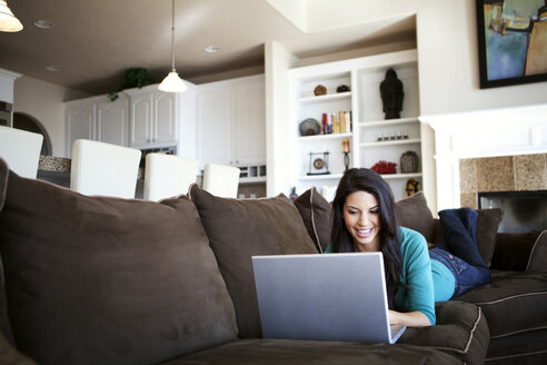 Junge Frau benutzt einen Laptop, während sie zu Hause auf dem Sofa liegt - CAVF00002