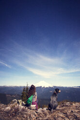 Rückansicht einer Frau, die mit ihrem Hund auf einer Klippe sitzt und auf einen Berg am Himmel blickt - CAVF00142