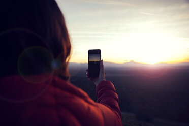Rückansicht einer Frau, die bei Sonnenuntergang mit ihrem Smartphone die Landschaft fotografiert - CAVF00146