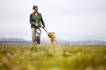 Happy woman walking with dog on grassy field against cloudy sky - CAVF00171