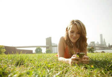 Smiling woman using smart phone while lying on grassy field against clear sky - CAVF00193