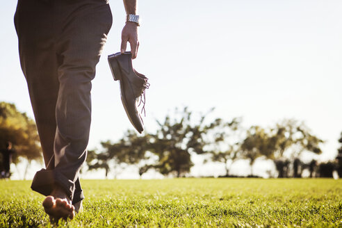 Low section of man holding shoes and walking on grassy field - CAVF00212