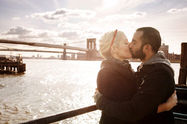 Paar, das sich küsst, während es an einem sonnigen Tag an der Brooklyn Bridge über den East River steht - CAVF00295