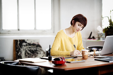 Businesswoman writing in book while working on laptop at desk in creative office - CAVF00311