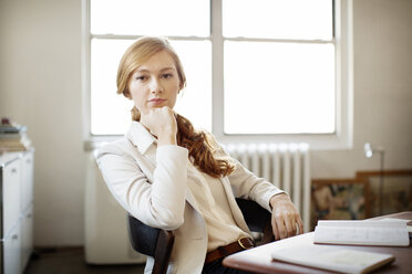 Portrait of confident businesswoman with hand on chin sitting at desk in office - CAVF00363