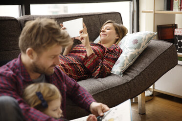 Vater und Tochter lesen ein Buch, während die Frau einen Tablet-Computer benutzt und sich auf der Couch entspannt - CAVF00419